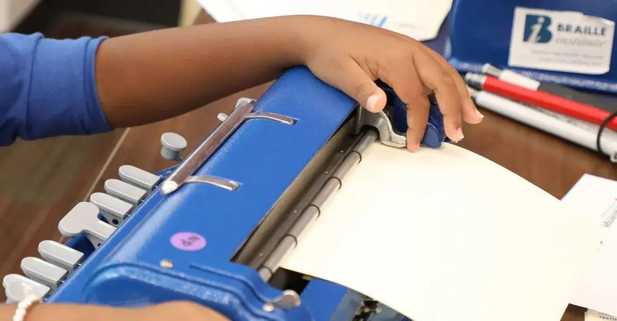 A child's hand operating a blue braille writer, feeding paper through the machine to emboss braille.