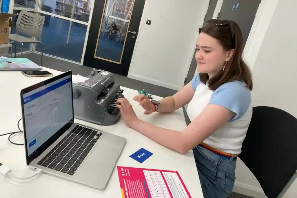 Seated at a white table inside a university group study room, Seren wears a smile while trying out Paige Connect with a braille writer and her laptop. The laptop screen showcases the Paige Connect web application in action.